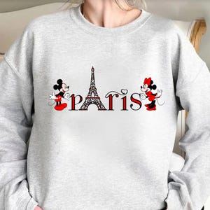 May include: A pale grey sweatshirt with the word "Paris" in red and black, incorporating the Eiffel Tower. Mickey and Minnie Mouse, in their iconic outfits, are positioned on either side of the word.