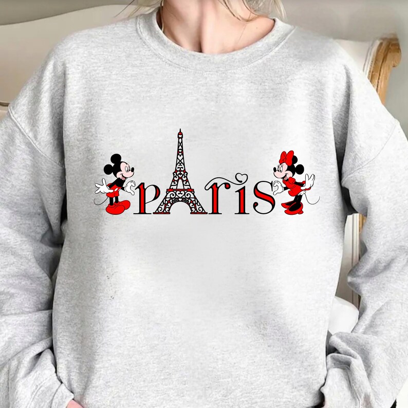 May include: A pale grey sweatshirt with the word "Paris" in red and black, incorporating the Eiffel Tower. Mickey and Minnie Mouse, in their iconic outfits, are positioned on either side of the word.