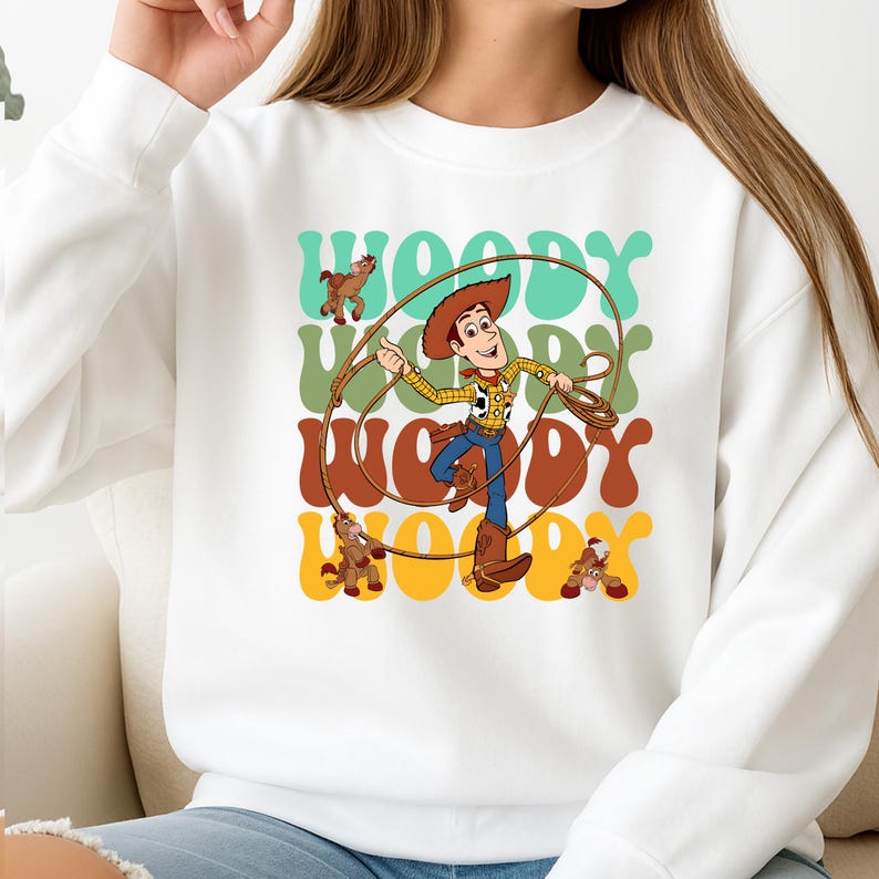 Puede incluir: Sudadera blanca con un gr&aacute;fico colorido del personaje Woody de Toy Story. El dise&ntilde;o incluye a Woody sosteniendo una cuerda, con la palabra "Woody" repetida en varios colores. Tambi&eacute;n se presentan caballos marrones.
