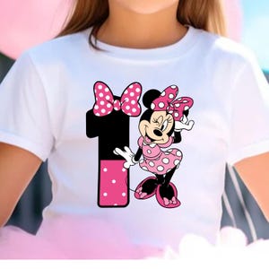 May include: White t-shirt featuring a graphic of Minnie Mouse and the number 1. Minnie Mouse is wearing a pink polka dot dress and bow. The number 1 is black with a pink polka dot section. A fun and festive design for a child's birthday.