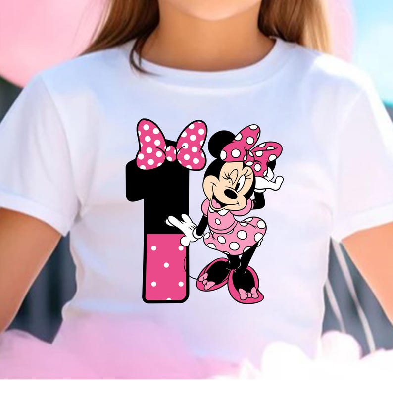 May include: White t-shirt featuring a graphic of Minnie Mouse and the number 1. Minnie Mouse is wearing a pink polka dot dress and bow. The number 1 is black with a pink polka dot section. A fun and festive design for a child's birthday.