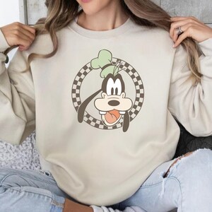 Op de afbeelding: Beige sweatshirt met een cartoonafbeelding van Goofy. Het ontwerp toont Goofy's gezicht in een bruin-witte geruite cirkel. Goofy draagt een groene hoed en steekt zijn tong uit. De sweatshirt heeft een neutrale kleur.
