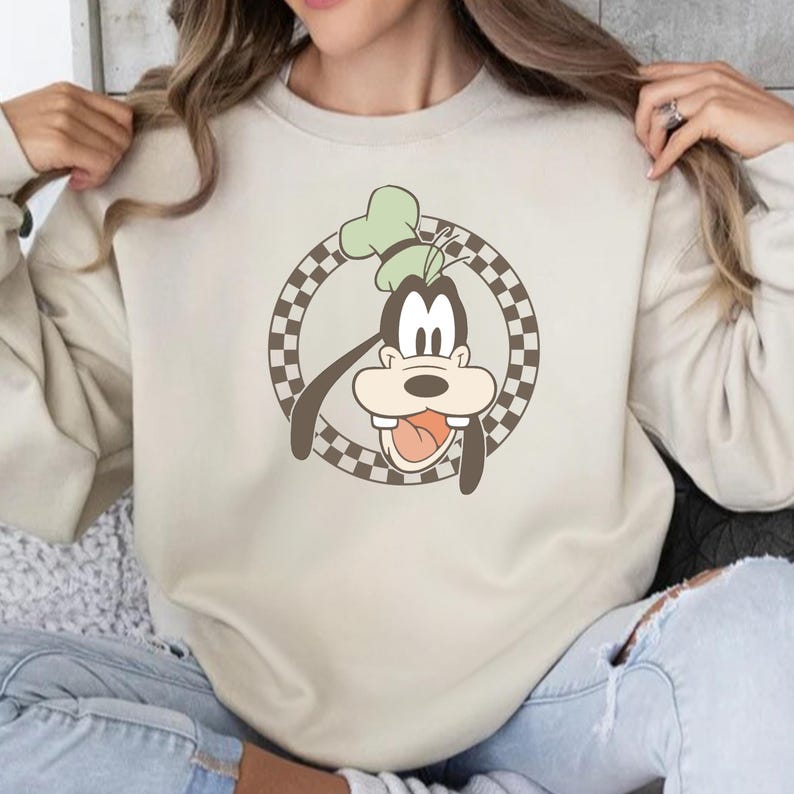 Op de afbeelding: Beige sweatshirt met een cartoonafbeelding van Goofy. Het ontwerp toont Goofy's gezicht in een bruin-witte geruite cirkel. Goofy draagt een groene hoed en steekt zijn tong uit. De sweatshirt heeft een neutrale kleur.