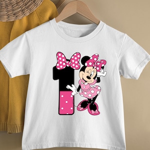 May include: White short-sleeved t-shirt featuring a graphic of Minnie Mouse in a pink polka-dot dress and bow. A large black number 1 with pink polka dots is also on the shirt. The shirt is hanging on a wooden hanger.