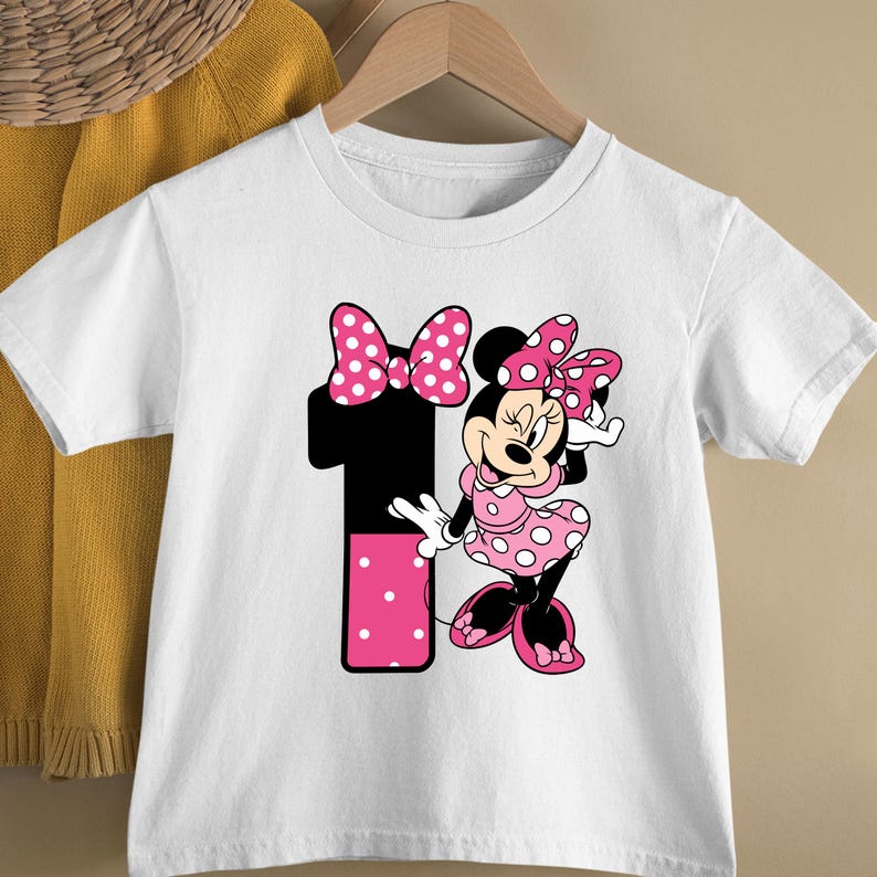 May include: White short-sleeved t-shirt featuring a graphic of Minnie Mouse in a pink polka-dot dress and bow. A large black number 1 with pink polka dots is also on the shirt. The shirt is hanging on a wooden hanger.