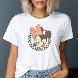 Op de afbeelding: Wit T-shirt met een Minnie Mouse-afbeelding, met een roze strik en een geruite cirkel. Het stripfiguur lacht. Het T-shirt is gemaakt van een zacht materiaal.