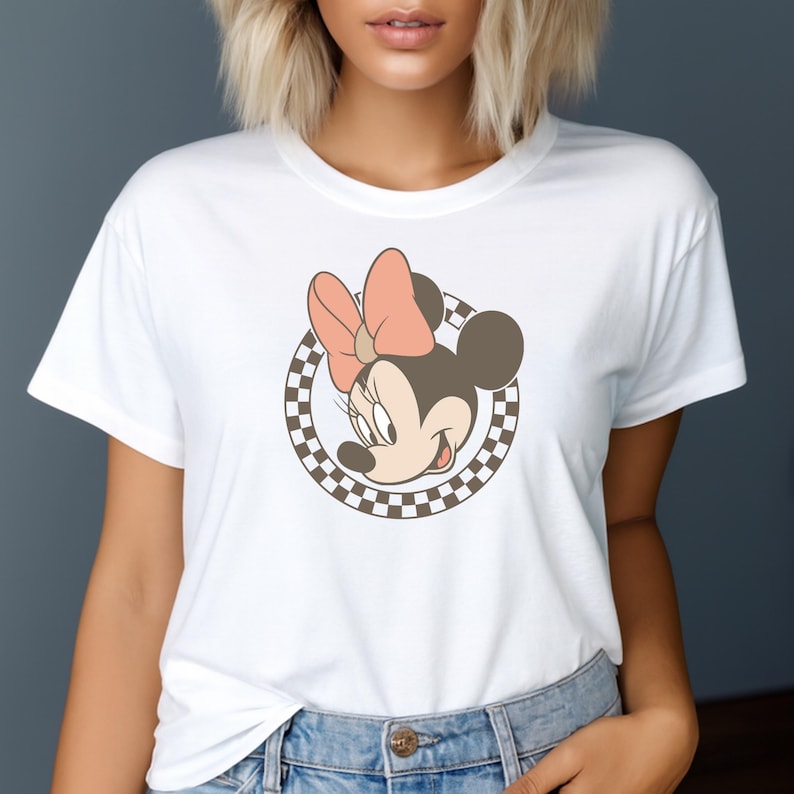 Op de afbeelding: Wit T-shirt met een Minnie Mouse-afbeelding, met een roze strik en een geruite cirkel. Het stripfiguur lacht. Het T-shirt is gemaakt van een zacht materiaal.