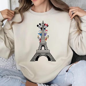 May include: A cream-coloured sweatshirt with a black Eiffel Tower design. The tower features Mickey Mouse, Minnie Mouse, Donald Duck, Daisy Duck, Goofy, and Pluto characters. The graphic is on the front of the long-sleeved top.