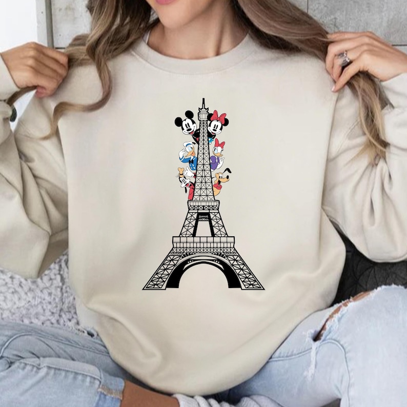 May include: A cream-coloured sweatshirt with a black Eiffel Tower design. The tower features Mickey Mouse, Minnie Mouse, Donald Duck, Daisy Duck, Goofy, and Pluto characters. The graphic is on the front of the long-sleeved top.