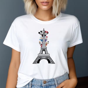 May include: White t-shirt with a black Eiffel Tower design and cartoon characters. The characters include Mickey Mouse, Minnie Mouse, Donald Duck, and Daisy Duck. The shirt has a crew neck.