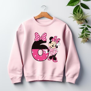 Puede incluir: Sudadera rosa claro con un gran n&uacute;mero 6 y un dise&ntilde;o de Minnie Mouse. El n&uacute;mero 6 es negro y rosa con lunares blancos. Minnie Mouse lleva un vestido y un lazo de lunares rosas. La sudadera est&aacute; colgada de una percha de madera.