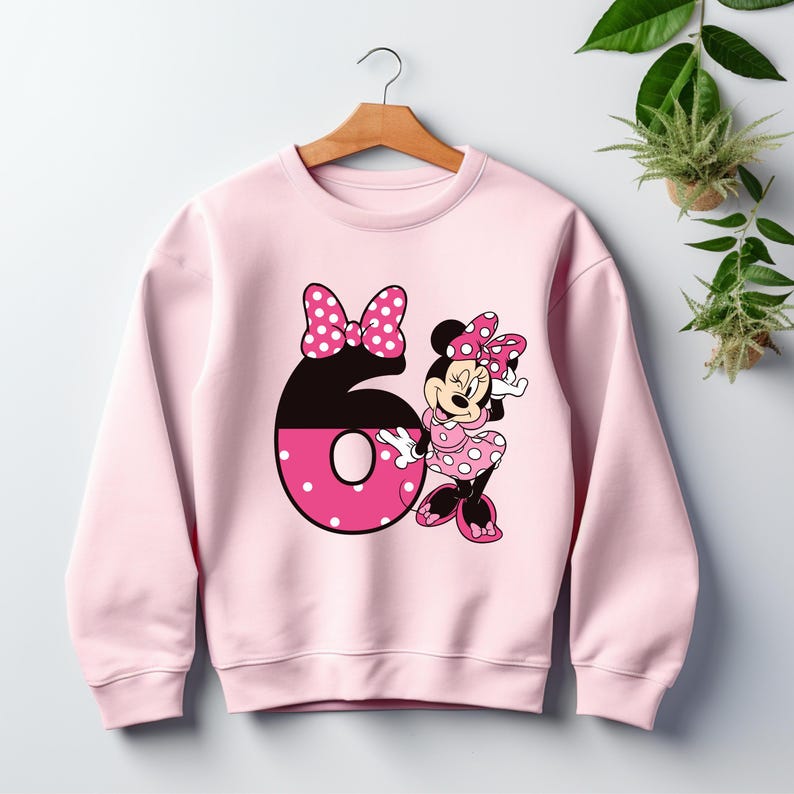 Puede incluir: Sudadera rosa claro con un gran n&uacute;mero 6 y un dise&ntilde;o de Minnie Mouse. El n&uacute;mero 6 es negro y rosa con lunares blancos. Minnie Mouse lleva un vestido y un lazo de lunares rosas. La sudadera est&aacute; colgada de una percha de madera.