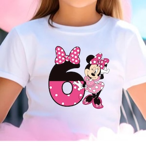 Puede incluir: Camiseta blanca con un gr&aacute;fico de Minnie Mouse y el n&uacute;mero 6. Minnie Mouse lleva un vestido y un lazo de lunares rosas. El n&uacute;mero 6 es negro y rosa con lunares blancos. Un lazo rosa est&aacute; encima del n&uacute;mero 6.