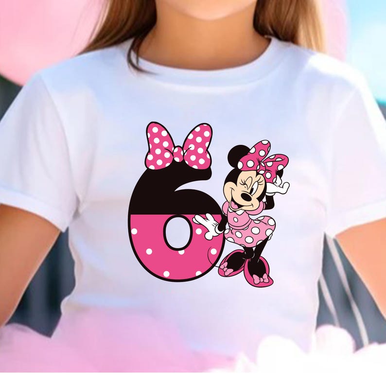 Puede incluir: Camiseta blanca con un gr&aacute;fico de Minnie Mouse y el n&uacute;mero 6. Minnie Mouse lleva un vestido y un lazo de lunares rosas. El n&uacute;mero 6 es negro y rosa con lunares blancos. Un lazo rosa est&aacute; encima del n&uacute;mero 6.