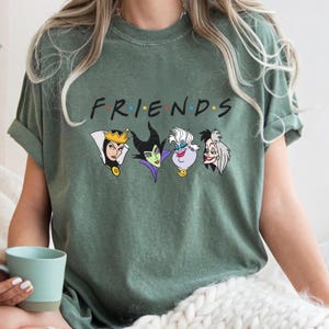 May include: Sage green t-shirt featuring the word "FRIENDS" in the style of the TV show logo, with the letters in different colors. Below the word are cartoon images of four Disney villains: The Evil Queen, Maleficent, Ursula, and Cruella de Vil.