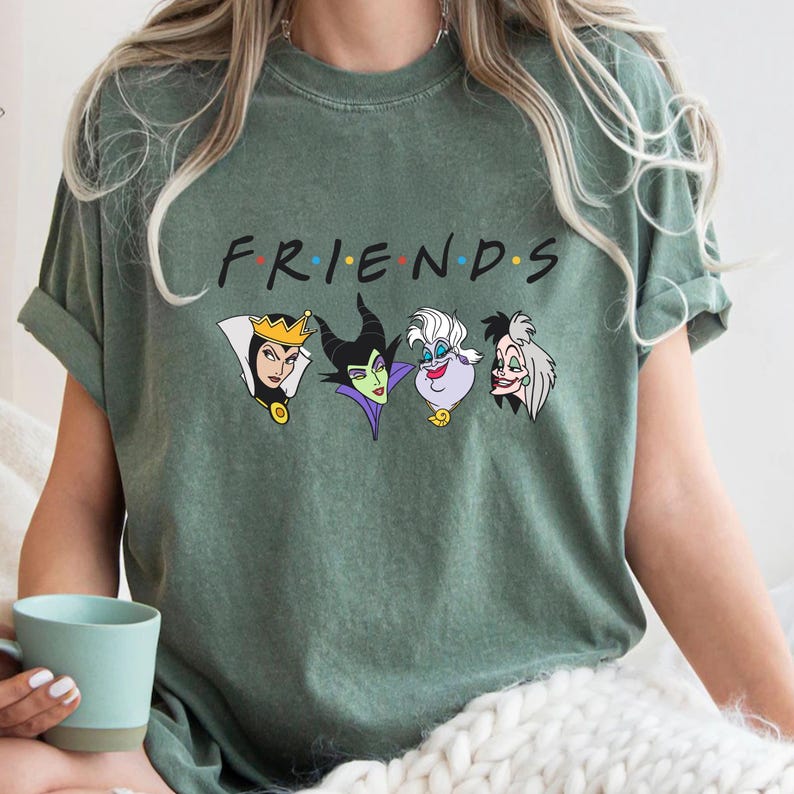 May include: Sage green t-shirt featuring the word "FRIENDS" in the style of the TV show logo, with the letters in different colors. Below the word are cartoon images of four Disney villains: The Evil Queen, Maleficent, Ursula, and Cruella de Vil.