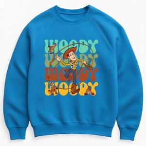 Puede incluir: Una sudadera azul con un gr&aacute;fico de Woody de Toy Story, sosteniendo un lazo. La palabra "WOODY" se repite en una fuente retro en tonos verdes, naranjas y marrones. El dise&ntilde;o incluye im&aacute;genes de dibujos animados de Woody y un caballo de juguete.
