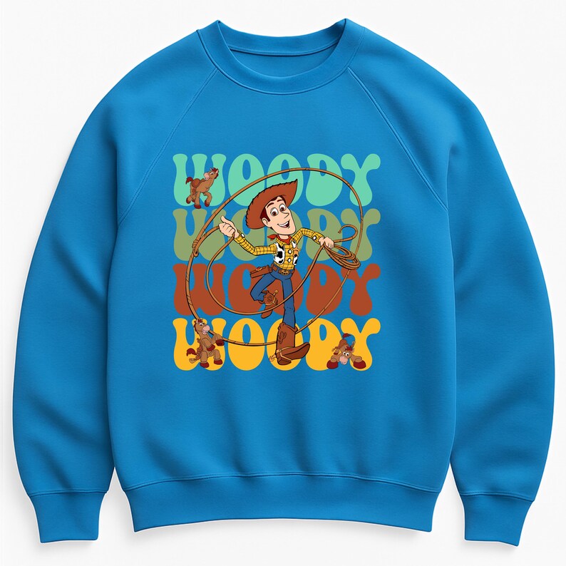 Puede incluir: Una sudadera azul con un gr&aacute;fico de Woody de Toy Story, sosteniendo un lazo. La palabra "WOODY" se repite en una fuente retro en tonos verdes, naranjas y marrones. El dise&ntilde;o incluye im&aacute;genes de dibujos animados de Woody y un caballo de juguete.