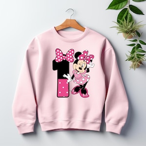 May include: A light pink sweatshirt featuring a graphic of Minnie Mouse and the number 1. Minnie Mouse is wearing a pink polka dot dress and bow. The number 1 is black with a pink polka dot design.