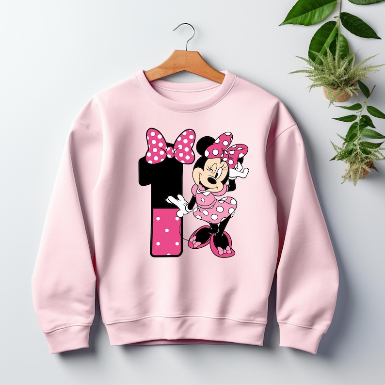 May include: A light pink sweatshirt featuring a graphic of Minnie Mouse and the number 1. Minnie Mouse is wearing a pink polka dot dress and bow. The number 1 is black with a pink polka dot design.
