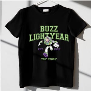 K&ouml;nnte beinhalten: Schwarzes T-Shirt mit einem gr&uuml;nen Aufdruck von Buzz Lightyear aus Toy Story. Das Design enth&auml;lt den Text "BUZZ LIGHTYEAR" mit "EST. 1995" und "TOY STORY" unter der Charakterillustration. Das T-Shirt h&auml;ngt an einem wei&szlig;en Kleiderb&uuml;gel.