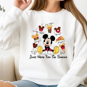 Puede incluir: Sudadera blanca con Mickey Mouse y varias ilustraciones de comida, como pizza y helado. El texto "Just Here For The Snacks" est&aacute; impreso debajo. El dise&ntilde;o es colorido y divertido.