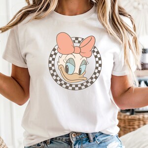 Op de afbeelding: Wit T-shirt met een cartoonafbeelding van Daisy Duck. De afbeelding toont een perzikkleurige strik, lichtblauwe ogen en een bruine snavel, omlijst door een zwart-wit geruite cirkel. Het shirt heeft korte mouwen en een ronde hals.