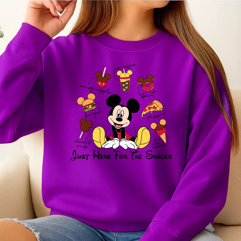 Puede incluir: Una sudadera morada con Mickey Mouse rodeado de iconos de comida como pizza y helado. El texto dice "Just Here For The Snacks". El dise&ntilde;o es de estilo caricatura.