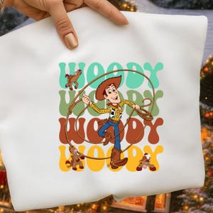 Puede incluir: Bolso tote blanco con una ilustraci&oacute;n de dibujos animados de Woody de Toy Story. El dise&ntilde;o incluye a Woody con sombrero y atuendo de vaquero, sosteniendo un lazo, con la palabra "WOODY" en una fuente retro. Tambi&eacute;n se incluyen peque&ntilde;as ilustraciones de caballos.