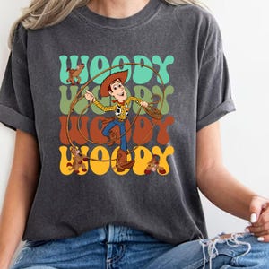 Puede incluir: Camiseta gris oscuro con una ilustraci&oacute;n de dibujos animados de Woody de Toy Story. El dise&ntilde;o incluye la palabra "Woody" en letras retro en verde azulado, verde, naranja y amarillo. Woody sostiene una cuerda.
