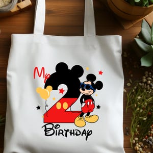 May include: White tote bag featuring a colorful Mickey Mouse design for a second birthday. The design includes a large number 2, Mickey Mouse wearing sunglasses, the words "My Birthday", and star and balloon accents.