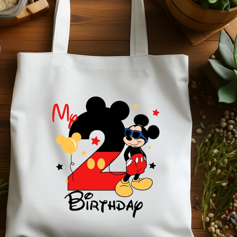 May include: White tote bag featuring a colorful Mickey Mouse design for a second birthday. The design includes a large number 2, Mickey Mouse wearing sunglasses, the words "My Birthday", and star and balloon accents.