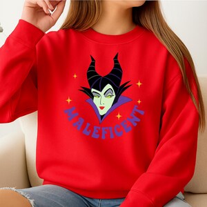 Puede incluir: Una sudadera roja con un gr&aacute;fico de la cara de Mal&eacute;fica. El dise&ntilde;o incluye cuernos negros, piel verde y un cuello morado. La palabra "MALEFICENT" est&aacute; escrita en azul alrededor de la imagen, con estrellas amarillas.