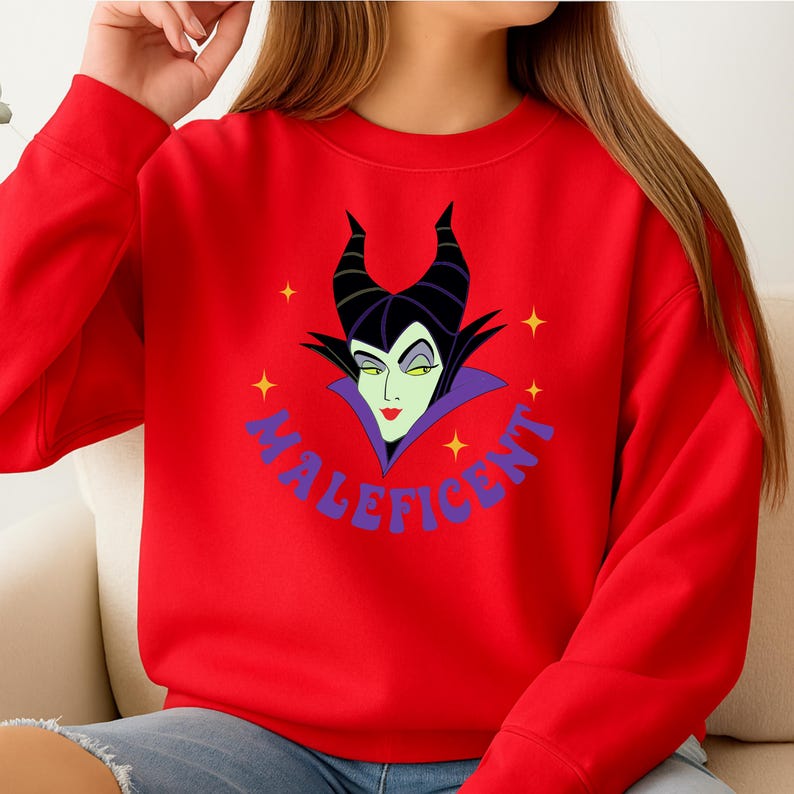 Puede incluir: Una sudadera roja con un gr&aacute;fico de la cara de Mal&eacute;fica. El dise&ntilde;o incluye cuernos negros, piel verde y un cuello morado. La palabra "MALEFICENT" est&aacute; escrita en azul alrededor de la imagen, con estrellas amarillas.