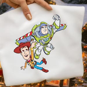 K&ouml;nnte beinhalten: Wei&szlig;er Kissenbezug mit einer farbenfrohen Grafik von Buzz Lightyear und Woody aus Toy Story. Buzz Lightyear tr&auml;gt einen gr&uuml;nen und wei&szlig;en Raumanzug, und Woody tr&auml;gt einen Cowboyhut und eine Weste.