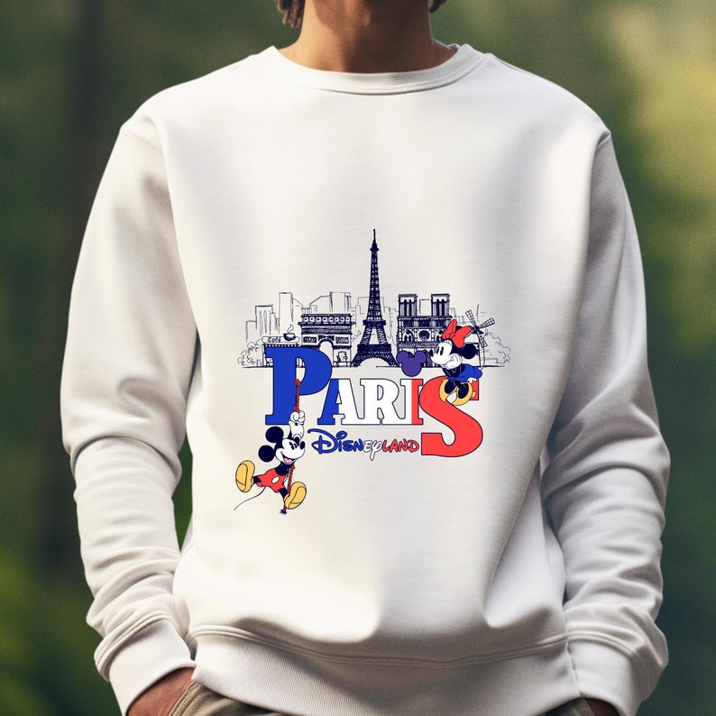 May include: White sweatshirt with a graphic of the Paris skyline, the Eiffel Tower, and Mickey and Minnie Mouse. The word "PARIS" is in blue and red, with "Disneyland" underneath. A casual top.