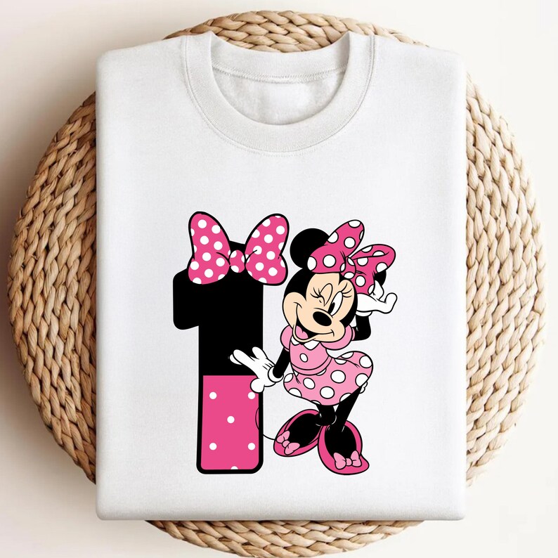 May include: White sweatshirt featuring a graphic of Minnie Mouse in a pink polka dot dress, holding a matching bow. A large black number 1 with pink polka dots and a pink bow is also on the sweatshirt.