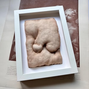 May include: A framed, beige-colored sculpture with rounded forms, resembling a body part. The artwork is presented in a white frame, set against a backdrop of vintage papers and illustrations. The sculpture's texture and shape are the focal points.