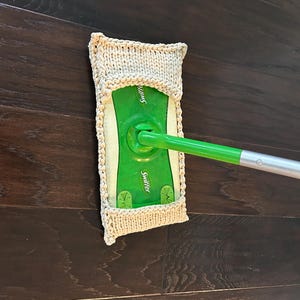 May include: A green and white Swiffer mop head with a beige knitted cover. The mop has a green handle and is positioned on a dark brown wooden floor. The knitted cover is rectangular and fits over the mop head.