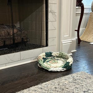 May include: A circular pet bed made of fabric scraps in shades of green, white, and pink sits on a dark wood floor. The bed is near a fireplace with a white marble tile surround and a gray rug.