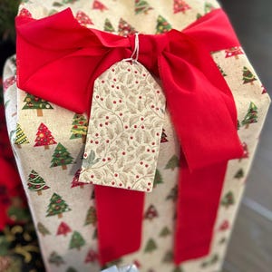May include: A wrapped gift box with a festive Christmas theme. The box is wrapped in cream-colored paper printed with colorful Christmas trees. A large red ribbon is tied around the box, and a decorative tag hangs from the ribbon.