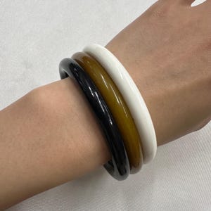 Puede incluir: Un conjunto de tres pulseras tipo brazalete lisas y redondeadas que se llevan juntas. El conjunto consta de un brazalete negro sólido, uno marrón ámbar translúcido y uno blanco sólido, todos con un diseño circular simple y pulido.
