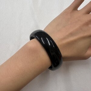 May include: A polished, black bangle bracelet. The bracelet is a solid, rounded band, reflecting light. The jewellery is worn on a wrist, set against a white background. The bracelet appears to be made of a smooth, polished material.