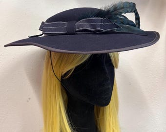 Puede incluir: Un sombrero azul marino de ala ancha con una banda de tela a juego que presenta finas rayas horizontales blancas. El sombrero está adornado con una pluma oscura e iridiscente insertada en el lateral de la banda, creando un aspecto estructurado.