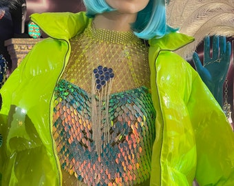 May include: A lime green puffer jacket with a zipper closure. The jacket is worn over a iridescent sequined top with a mesh overlay. The model is wearing a silver sequined baseball cap and blue jeans.