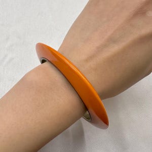 Op de afbeelding: Een massieve, gladde, feloranje armband met een afgerond, dik profiel. De armband heeft een glanzende afwerking en een eenvoudig, doorlopend cirkelvormig ontwerp dat stevig om de pols zit en een levendige en gedurfde kleur laat zien.