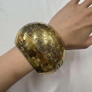 Op de afbeelding: Een brede, goudkleurige manchetarmband met een geëtst bloemenontwerp en een gehamerde textuur. De armband is ongeveer 7.6 cm breed en wordt om een pols gedragen.