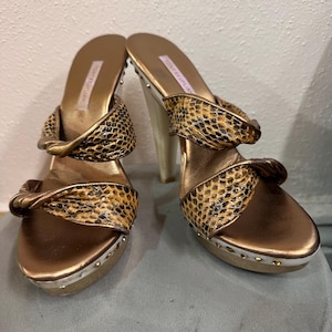 May include: A pair of gold-toned platform sandals with a snakeskin pattern on the straps. The sandals have a high heel and are embellished with small gold studs around the sole.