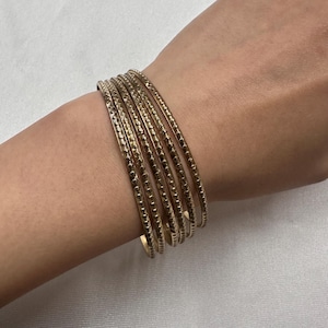 May include: A collection of six gold bangles. Each bangle features a textured surface with a repeating pattern of small, raised dots and lines. The bangles are stacked on a wrist, highlighting their design and colour.