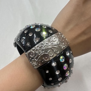 May include: A wide, black bangle bracelet with sparkling clear rhinestones and a silver-tone floral design. The bracelet is a bold statement piece, perfect for adding a touch of glamour to any outfit.
